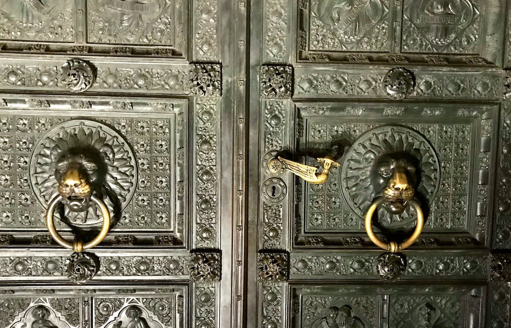 Περίτεχνες μεταλλικές πόρτες με περίτεχνα σχέδια, με χρυσά ρόπτρα κεφαλής λιονταριού και λεπτομερή χαρακτικά.