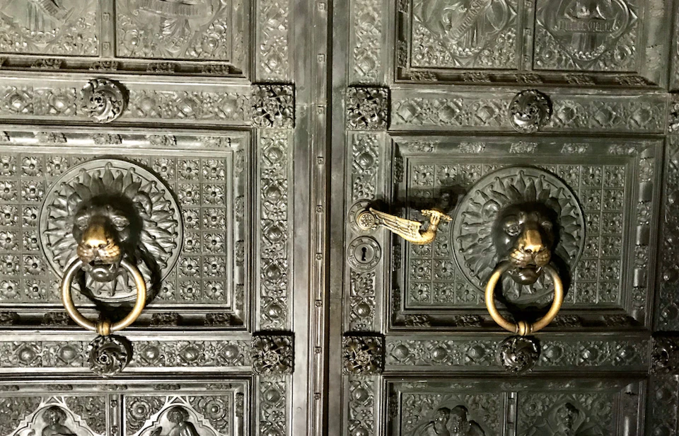 Ornate metal doors with intricate patterns, featuring golden lion head door knockers and detailed engravings.