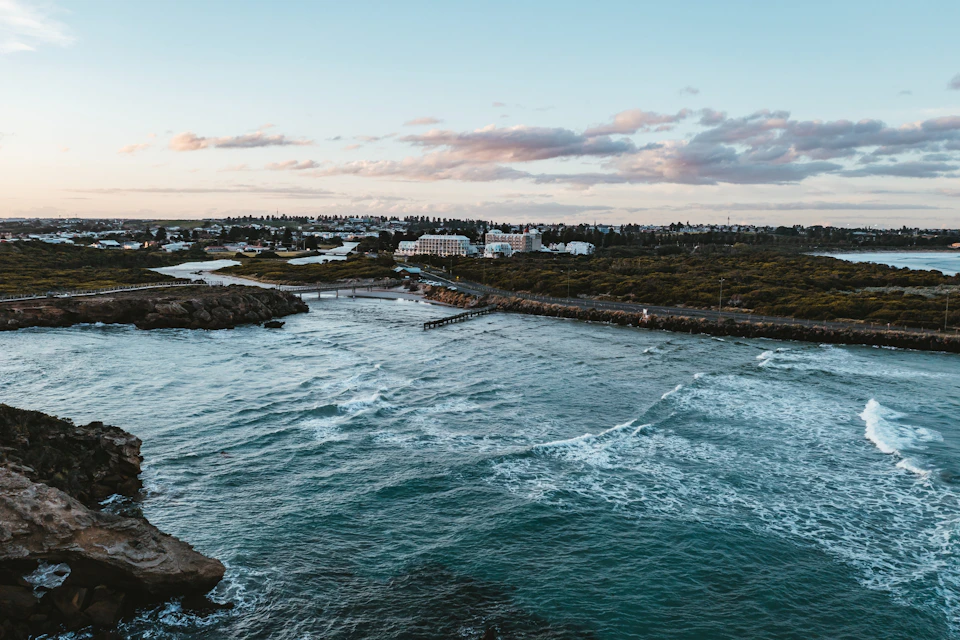 Coastal landscape with a calm sea, a small pier, and buildings surrounded by greenery under a partly cloudy sky.