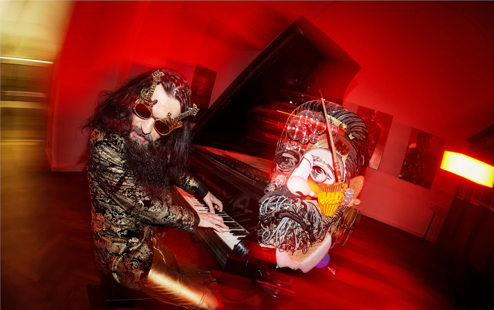 A person with long hair and sunglasses plays a piano next to a large, colorful, surreal sculpture of a face under red lighting.