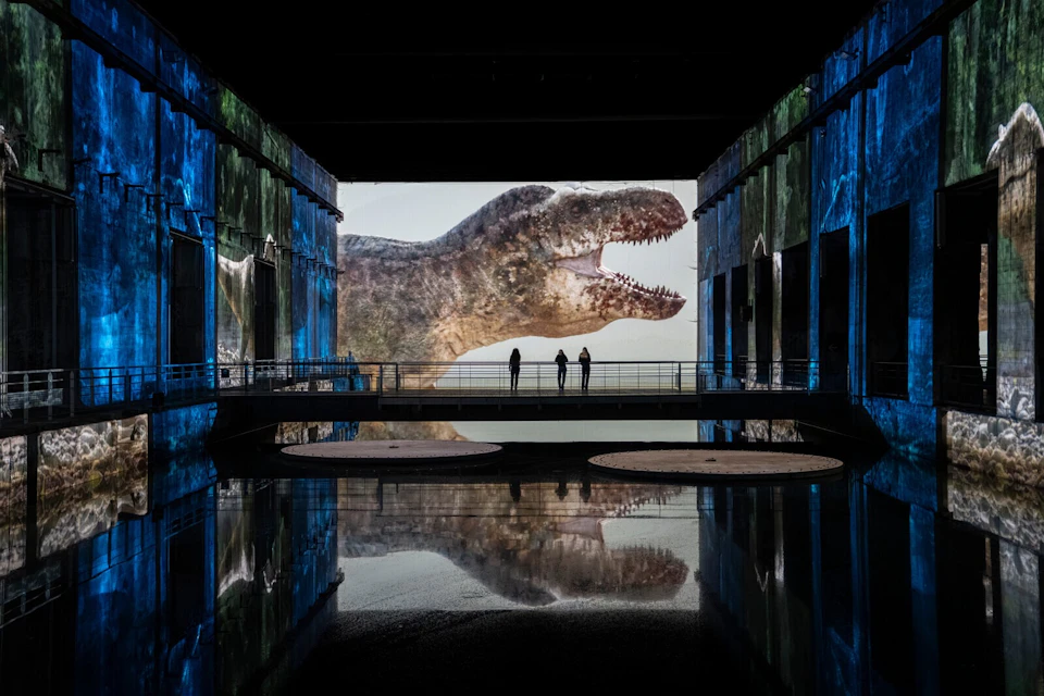 A large projection of a dinosaur head in a dark room with blue walls and three people standing on a bridge in the center.