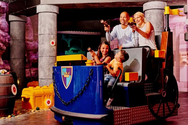 Una famiglia di quattro persone sale su un'attrazione del parco divertimenti, prendendo la mira con pistole giocattolo, sorridendo e godendosi l'esperienza.