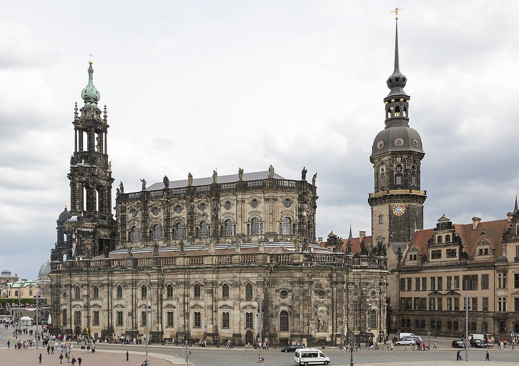 Historic stone cathedral with ornate facade and two tall towers, one with a clock. People and vehicles in the foreground. Cloudy sky.