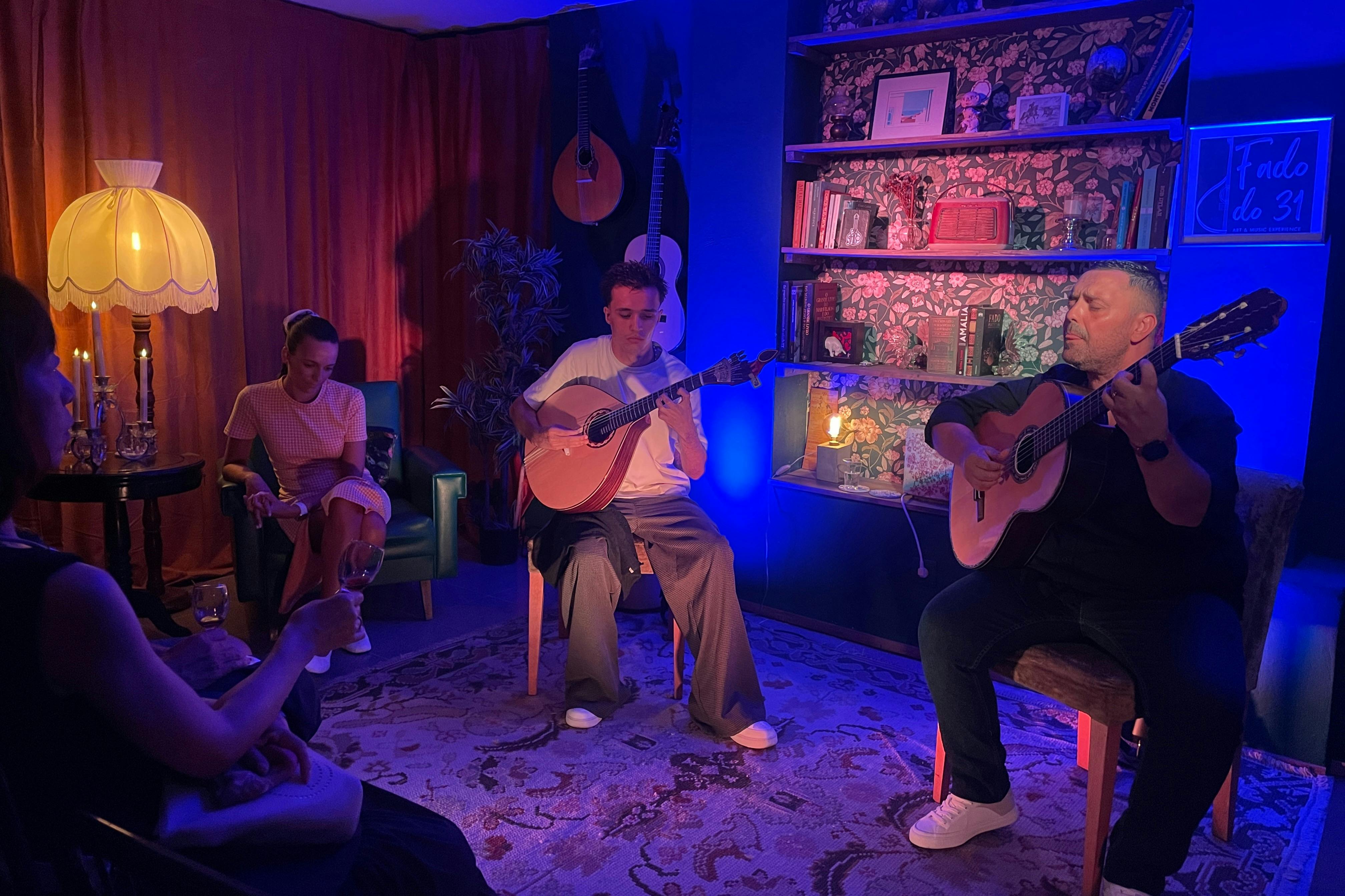 Δύο άνθρωποι παίζουν ακουστικές κιθάρες σε ένα άνετο δωμάτιο με φωτισμένα ράφια, βιβλία και διακοσμήσεις, κάτω από μπλε και μοβ φωτισμό.