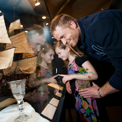 En man och en flicka tittar på en skeppsmodell i ett glasfodral på ett museum, ler och pekar på utställningen.