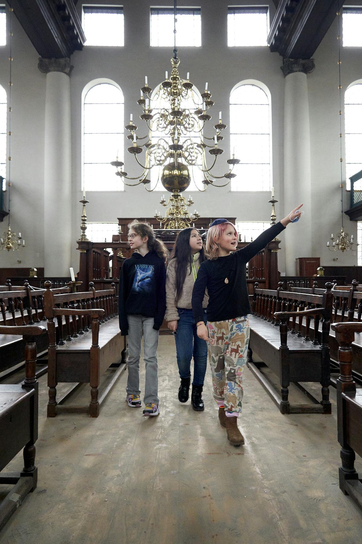Trois enfants marchent dans l'allée d'un grand hall avec de hautes fenêtres et des lustres ornés ; l'un d'entre eux pointe du doigt vers l'avant.