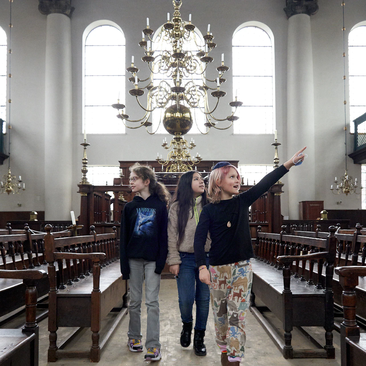 Three children walk down the aisle of a grand hall with tall windows and ornate chandeliers; one child points forward.