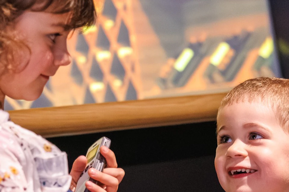 Two children holding glasses, standing indoors near a wooden counter, with wine racks visible in the background.