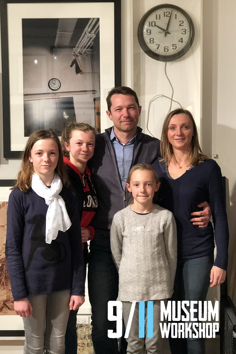A family of five stands together indoors, smiling. A clock and framed picture are visible on the wall behind them.