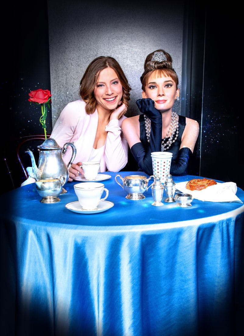 Two women sit at a blue-clothed table with a coffee set, a red rose, and a croissant. One wears a pearl necklace and black gloves.