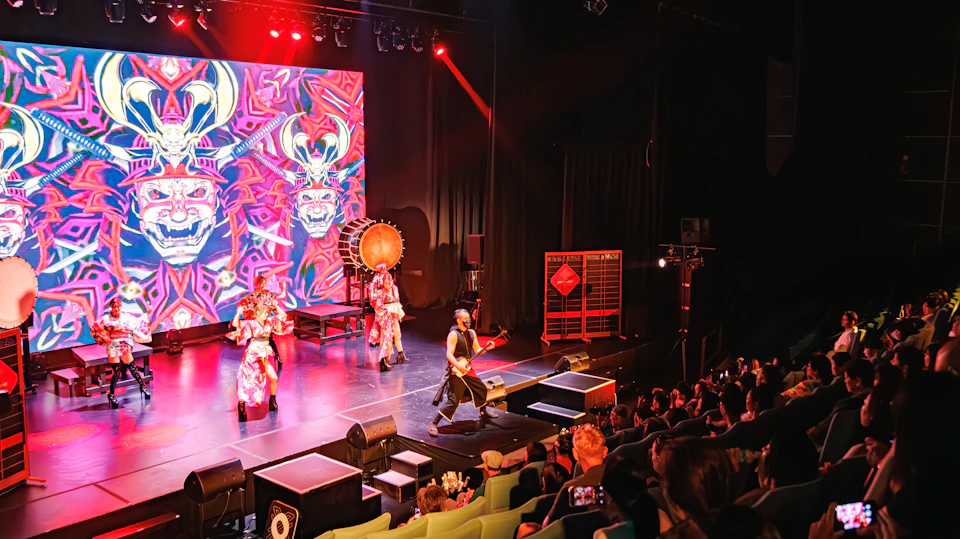 Performers in vibrant costumes on a stage with colorful visuals, a drum, and an audience seated in front.