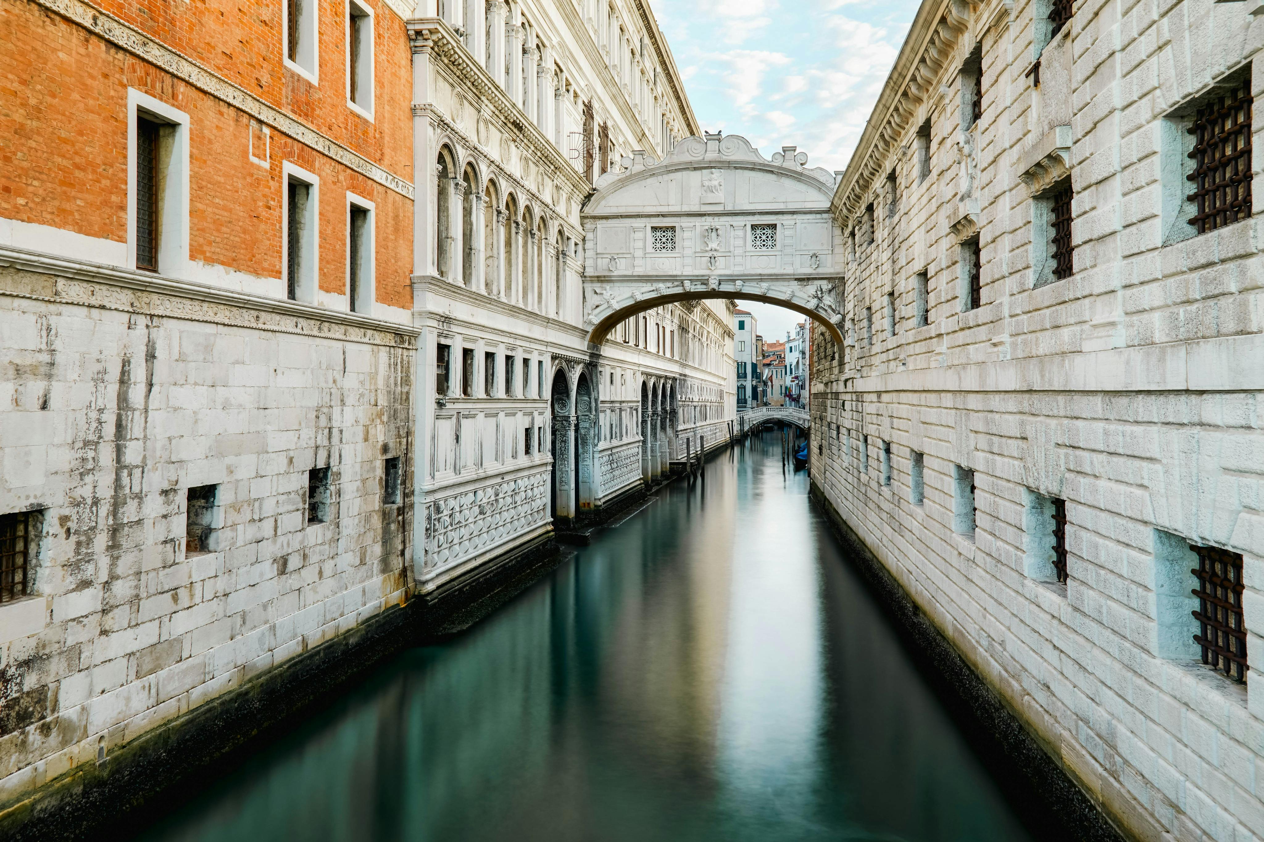 威尼斯狭窄的运河，两侧是带拱形窗户的历史建筑。 叹息桥（ Bridge of Sighs ）在部分多云的天空下横跨运河。