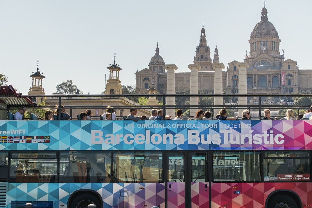 Offener Tour-Bus mit der Aufschrift "Barcelona Bus Turístic", gefüllt mit Menschen, vor der Kulisse historischer Gebäude und verschnörkelter Türme.