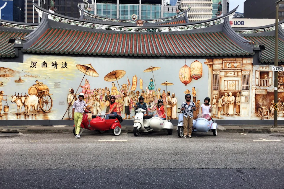 People with decorative scooters posing in front of a colorful mural depicting a historic scene, set against an urban backdrop.