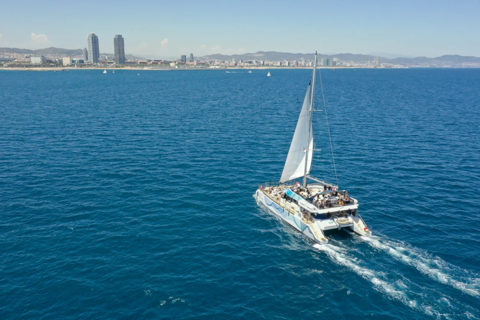 A catamaran sailing on a blue sea near a city with tall buildings and mountains in the background.