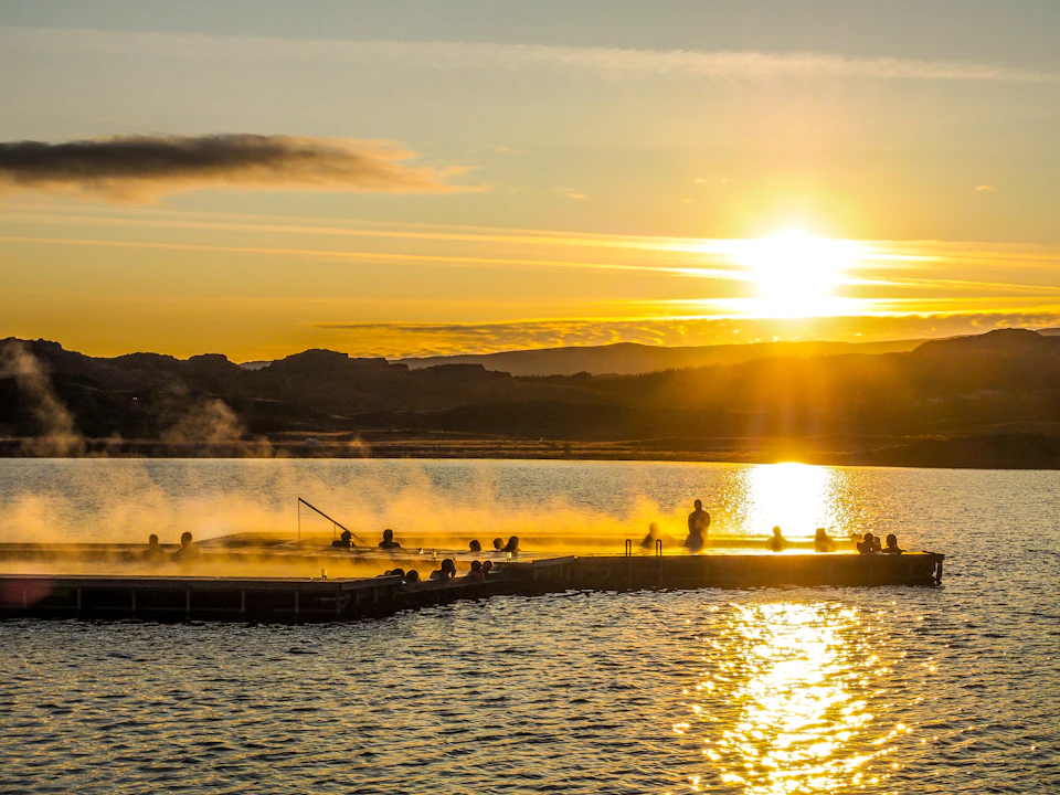 People relax in a steaming hot spring pool at sunset, with mountains and a lake in the background.