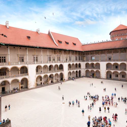 En innergård omgiven av flervåningsbyggnader med valv, röda tegeltak och grupper av människor som går eller samlas nedanför.
