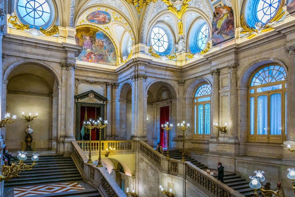 Interior of a grand hall with ornate architecture, arched windows, frescoes, and several staircases and chandeliers.
