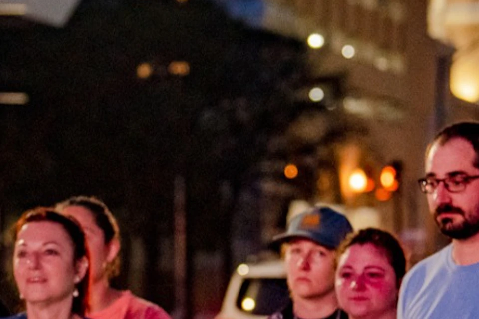 A group of people standing together outdoors at night in an urban setting with streetlights and buildings in the background.