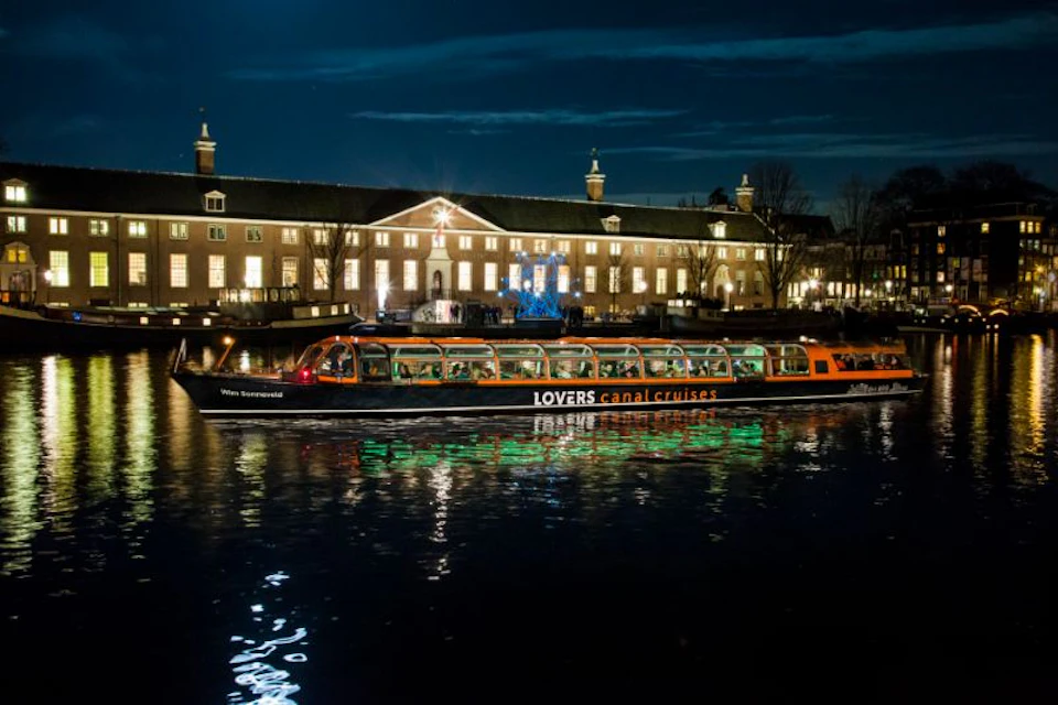 A illuminated canal cruise boat called "Lovers" passes by a lit-up building at night, reflecting light on the water.