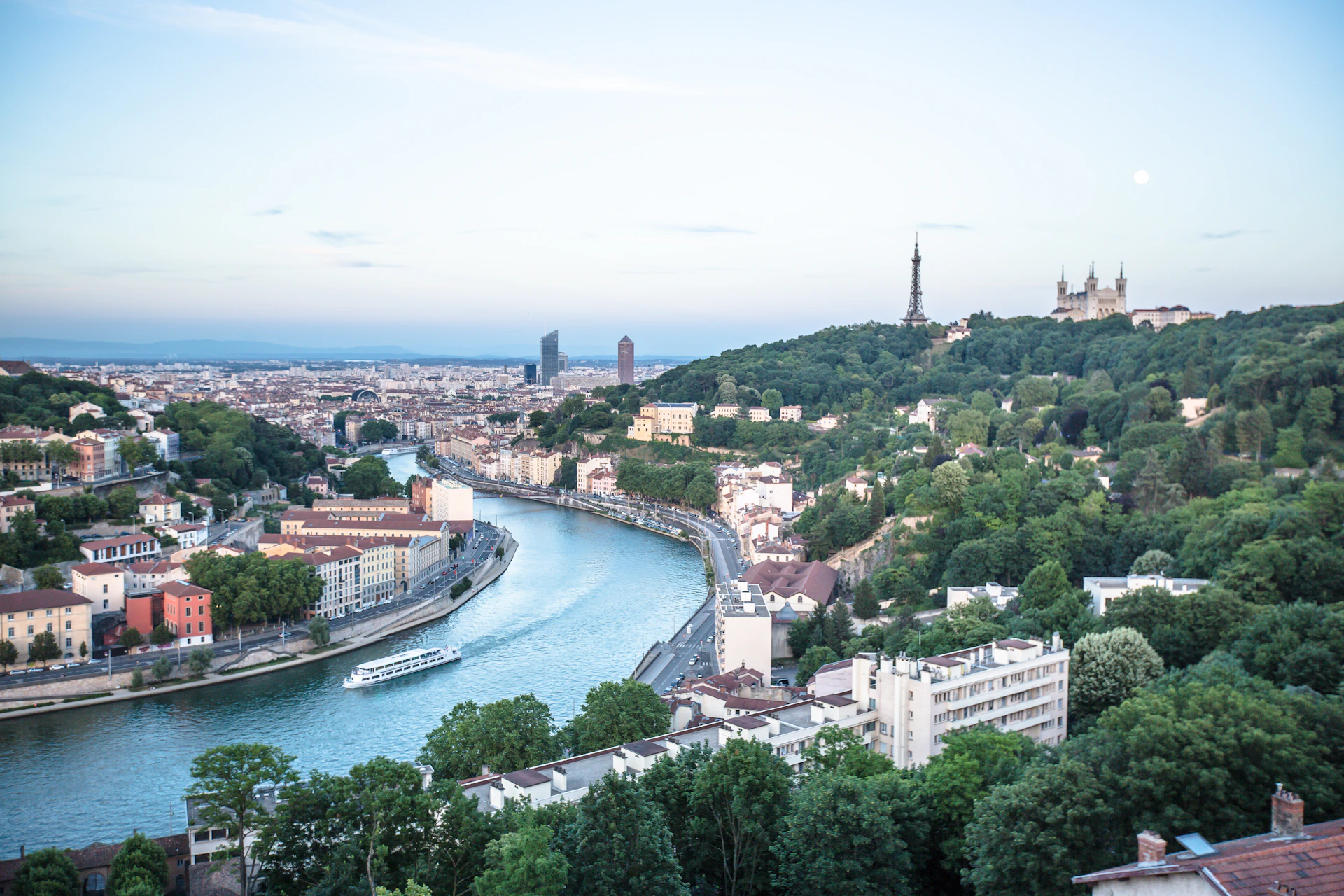 A cityscape featuring buildings along a river, a boat cruising on the water, and a hill with a tower and basilica in the distance.