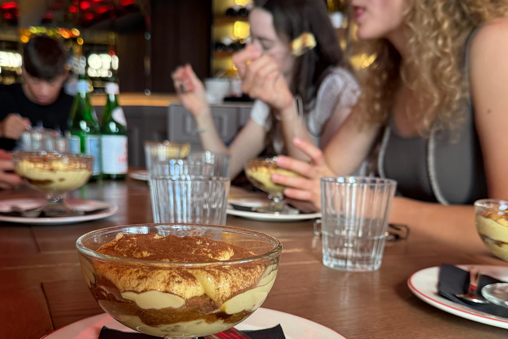 Two people enjoying dessert at a table with tiramisu in a glass bowl, water glasses, and bottles in the background.
