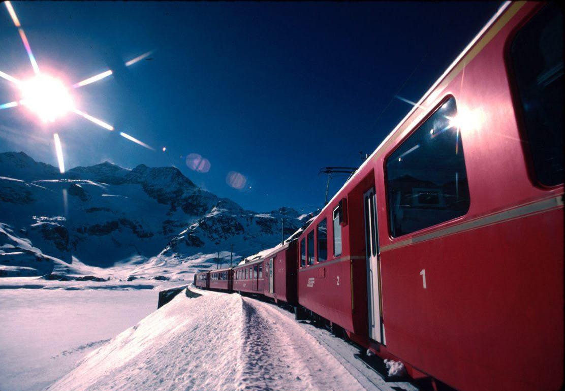 Ένα κόκκινο τρένο ταξιδεύει μέσα σε ένα χιονισμένο ορεινό τοπίο κάτω από έναν καταγάλανο ουρανό, αντανακλώντας το φως του ήλιου.