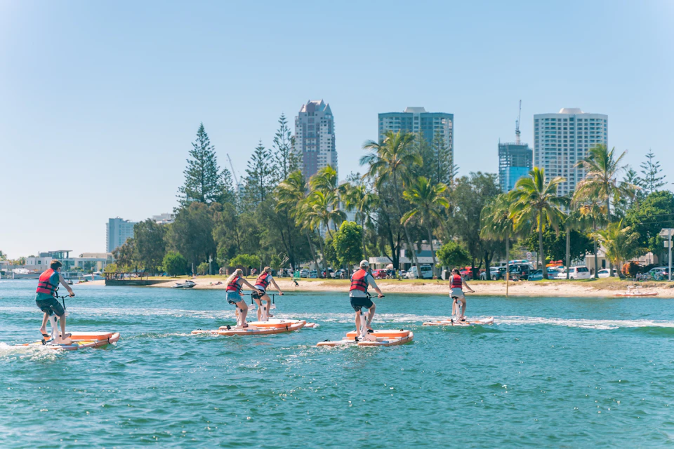 People riding large paddleboards on a calm body of water near a sandy shore with trees and tall buildings in the background.