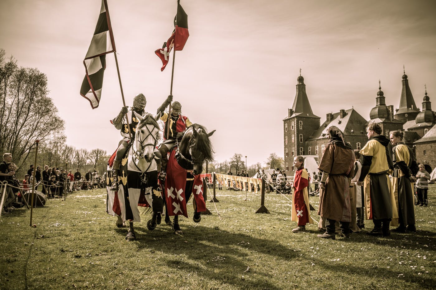 Two knights on horses, carrying flags, reenact a jousting scene at a medieval fair near a large castle. Spectators watch nearby.