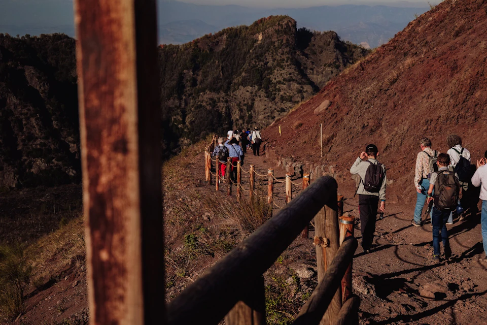 Hikers walk along a dirt trail bordered by wooden railings, with hills and trees in the background under a clear sky.