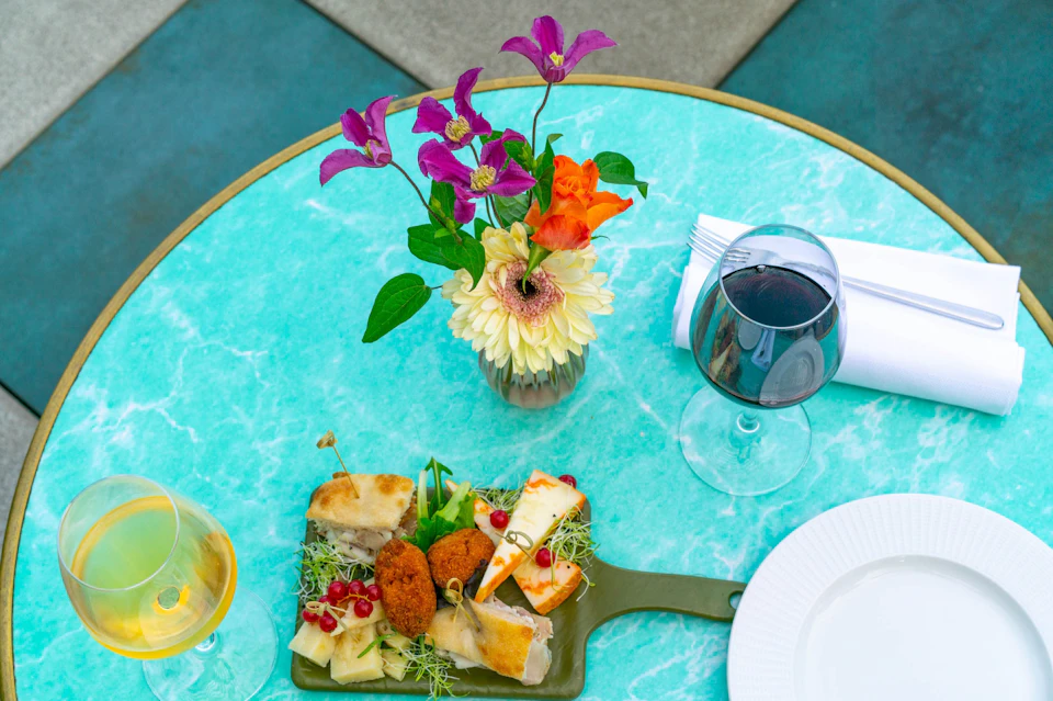 A colorful meal setup on a turquoise table, featuring a cheese platter, red wine, and a floral centerpiece in a vase.