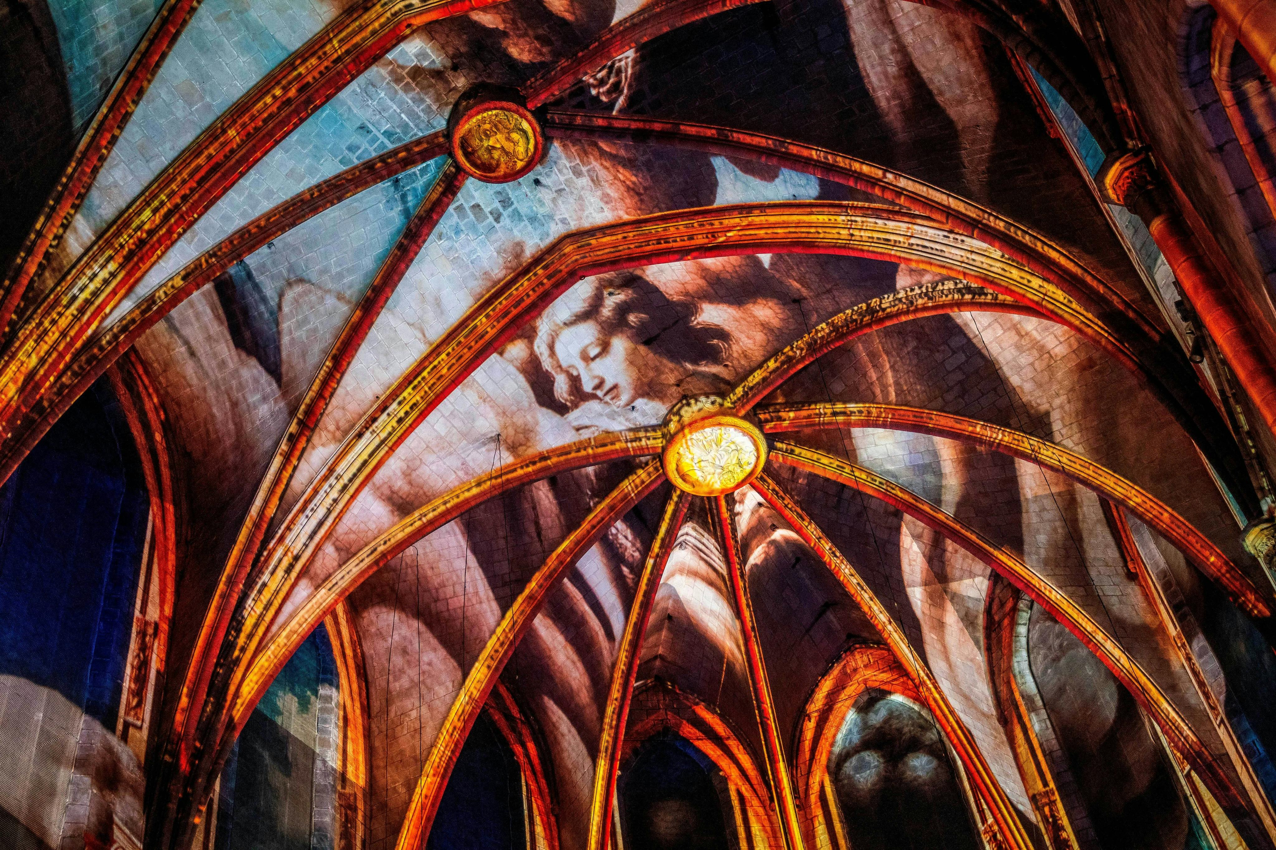 Un plafond de cathédrale avec des arcs complexes et une grande figure angélique peinte, éclairée par une lumière chaude.