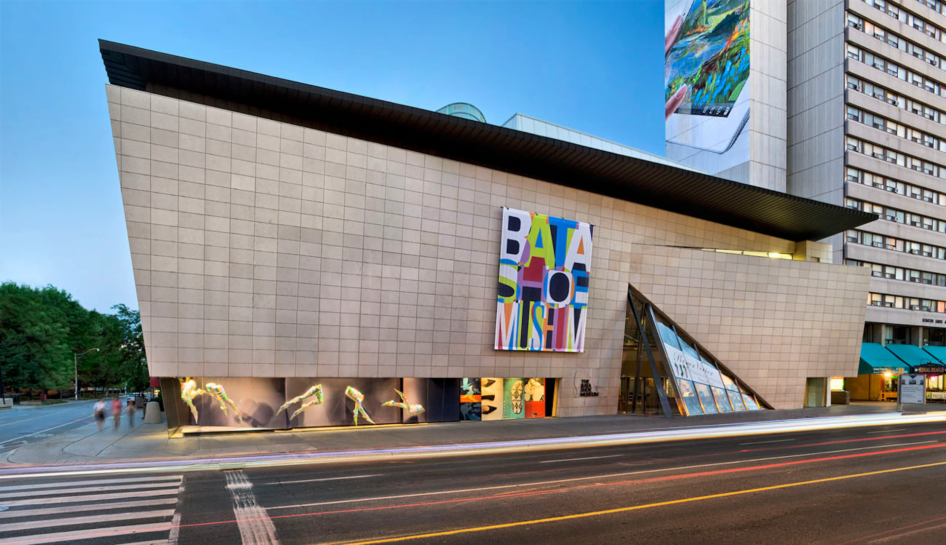 A beige building with a large multicolored "Bata Shoe Museum" sign, display windows, and a glass entrance beside a road.