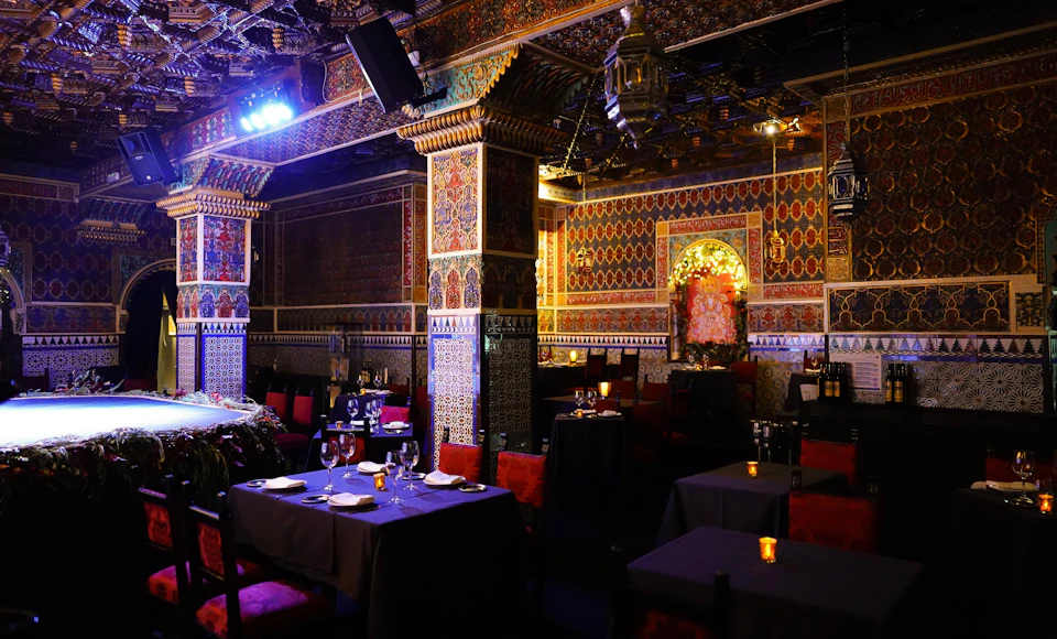 An ornate restaurant interior with colorful, intricate wall patterns, dim lighting, and tables set with wine glasses and candles.