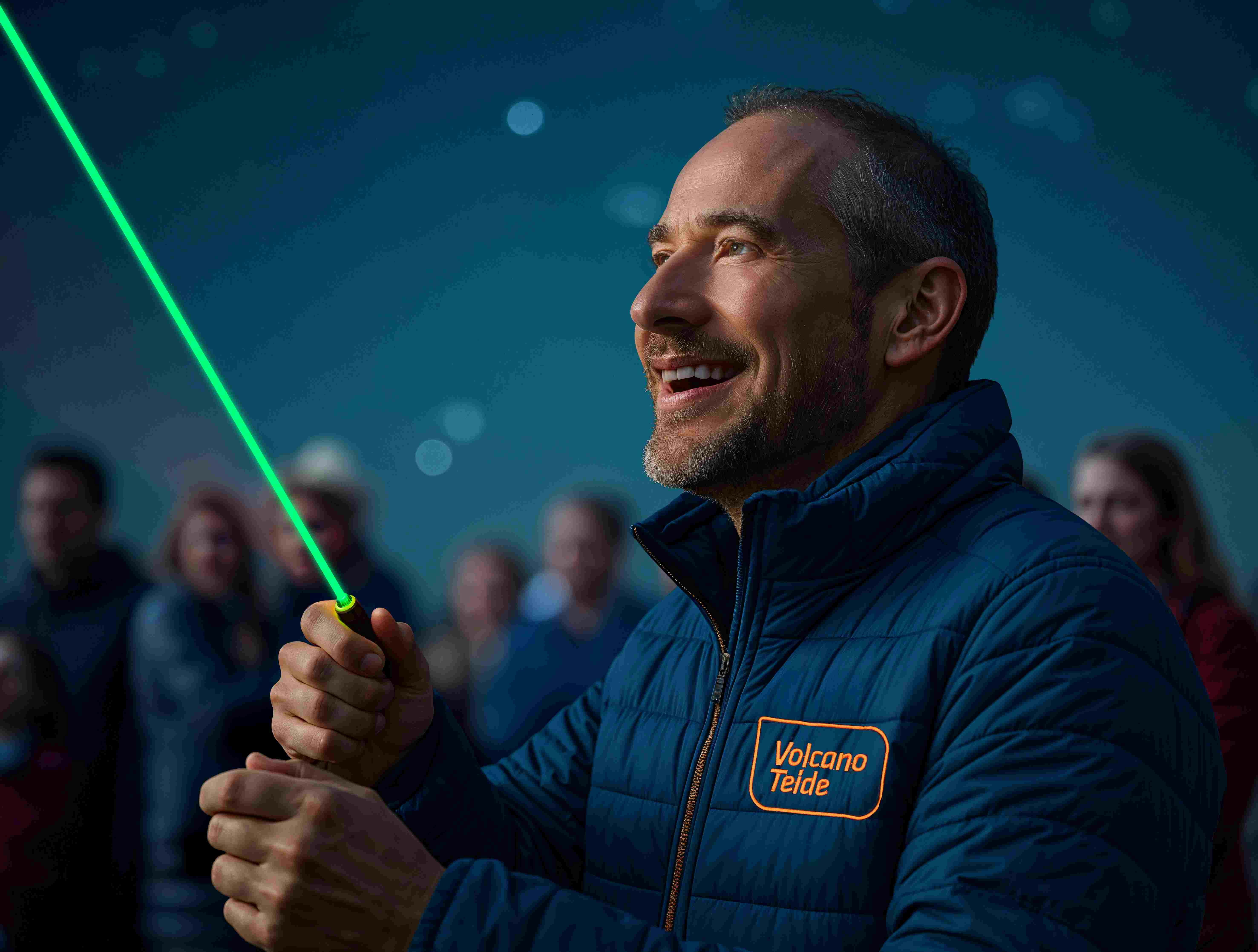 「ボルケーノ・テイデ」のロゴが入った青いジャケットを着た男性が、緑色のレーザーポインターを手にして微笑んでいます。背景にはぼやけた人々がいます。
