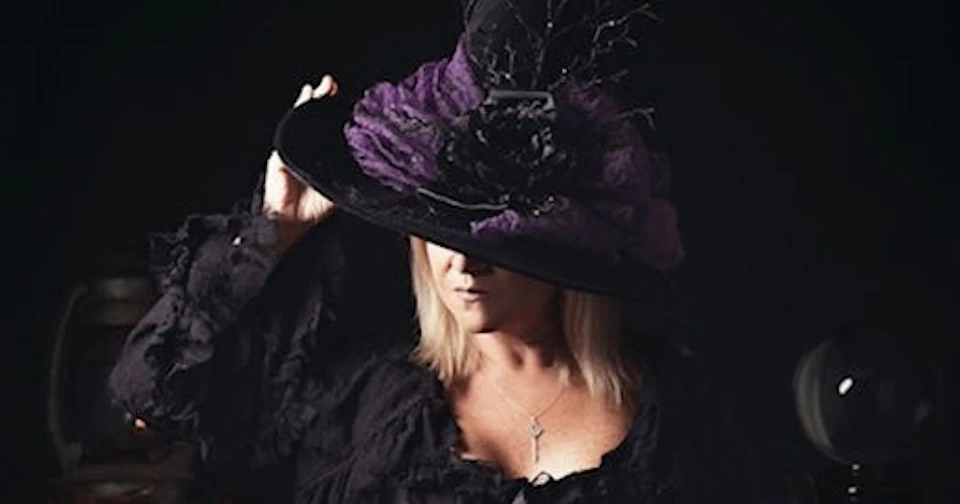 A person wearing a decorative black and purple hat with a feather, dressed in black and posing against a dark background.