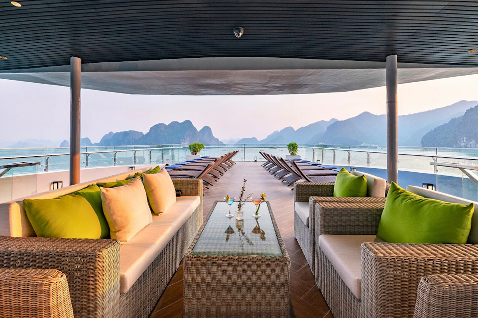 An outdoor lounge area with wicker sofas and green pillows overlooks lounge chairs, a glass railing, and a mountainous bay.