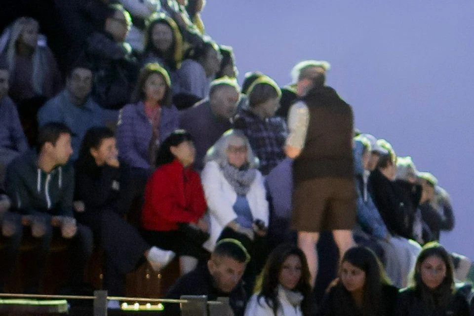 A crowd of people watches penguins on a sandy beach, with wooden seating and a glass viewing area, during dusk.