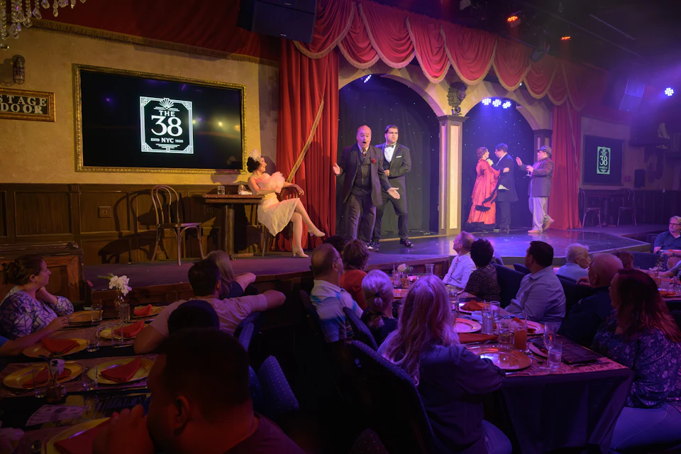 A stage performance in progress with actors in period costumes and a seated audience dining at tables in a theater.