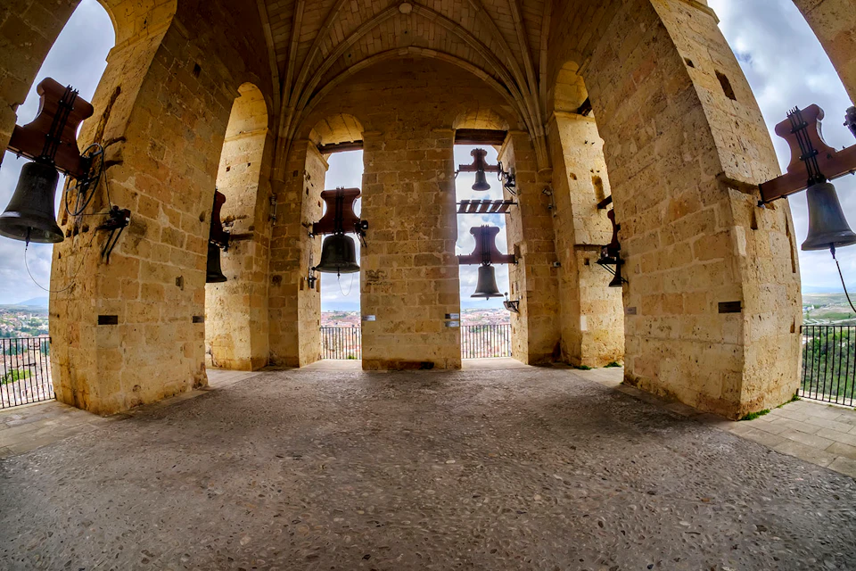 An interior view of a stone bell tower with several large bells hanging in the archways. A cityscape is visible through the openings.