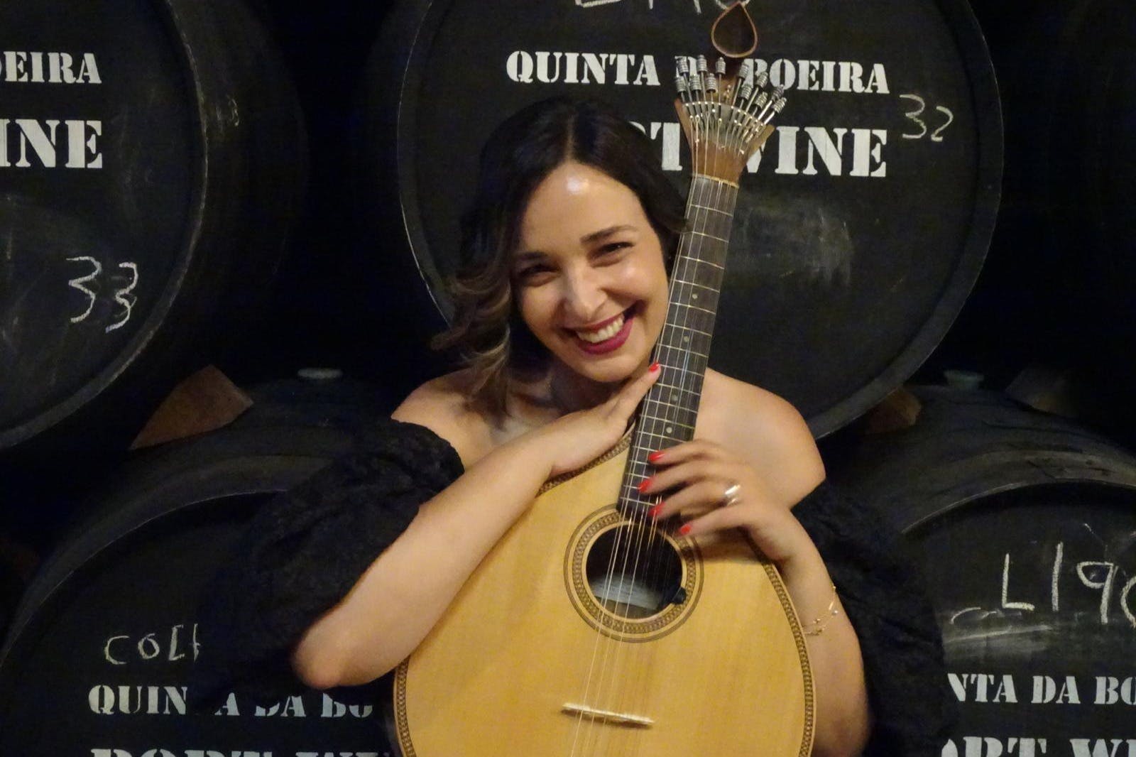 Usmívající se žena držící portugalskou kytaru stojí před velkými sudy s vínem označenými „PORTSKÉ VÍNO“ a „QUINTA DA BOEIRA“.