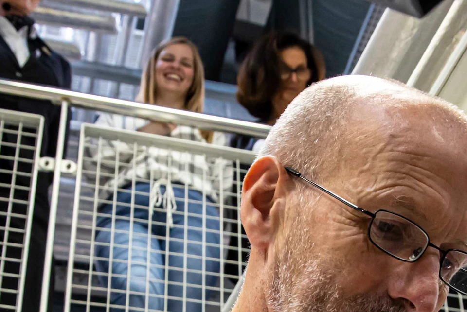 Bald man in a white coat works with machinery, while a group observes from a balcony above.