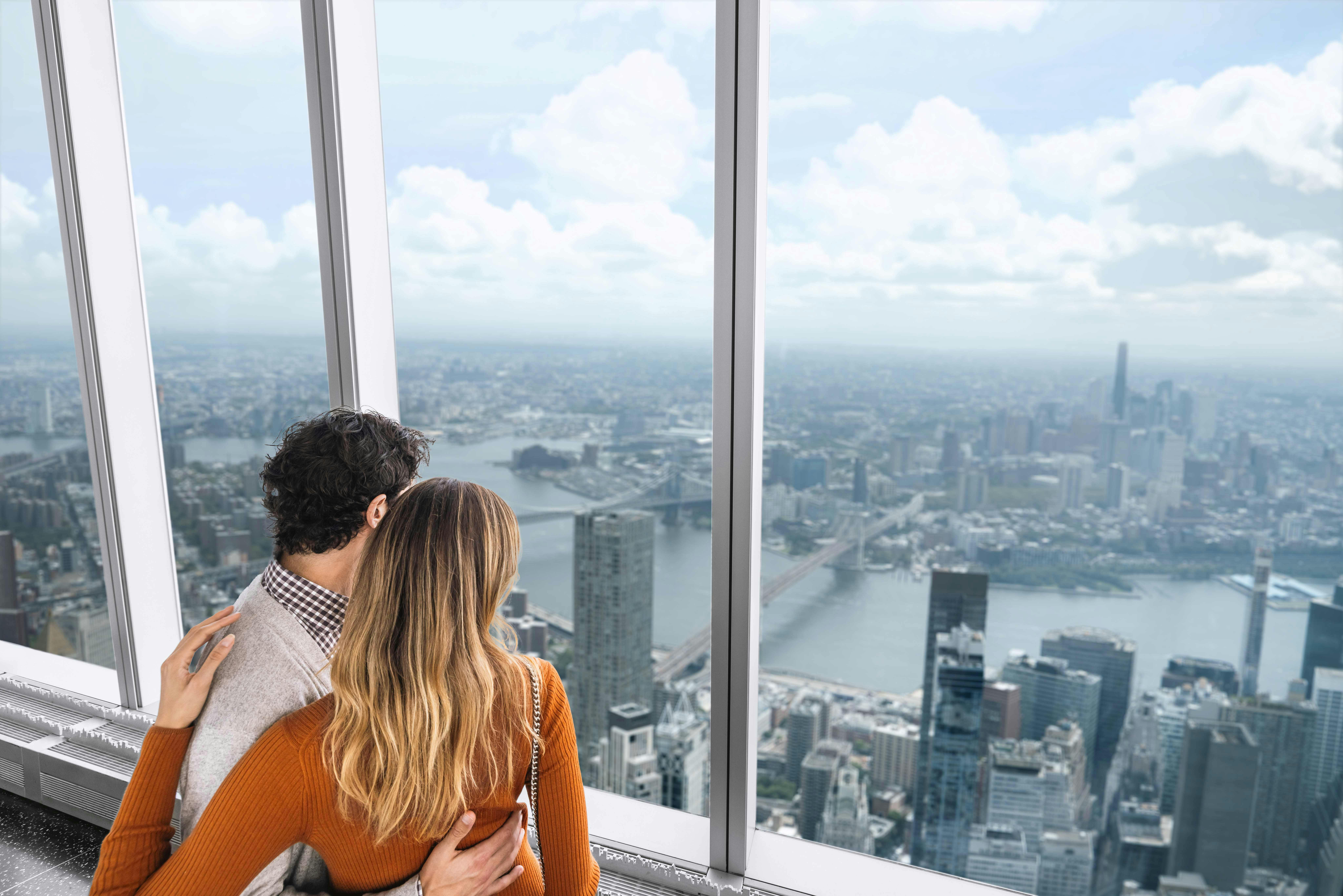A couple embraces while looking out from a high observation deck window, overlooking a sprawling cityscape with rivers and skyscrapers.