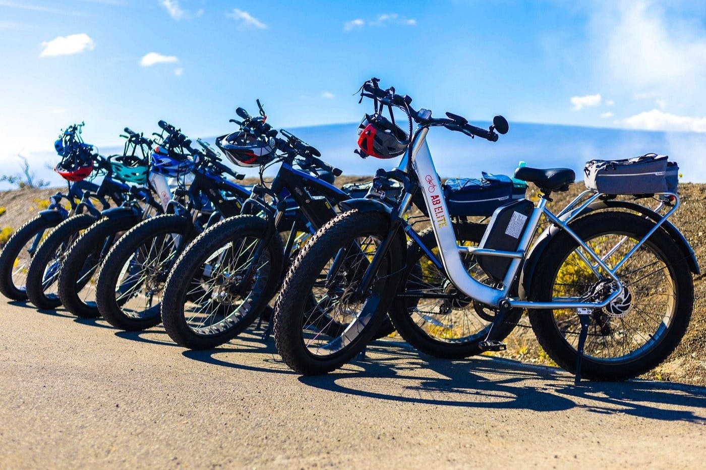 포장된 표면에 7대의 자전거가 줄지어 주차되어 있고, 핸들바에 헬멧이 매달려 있으며, 맑고 푸른 하늘 아래 있습니다.