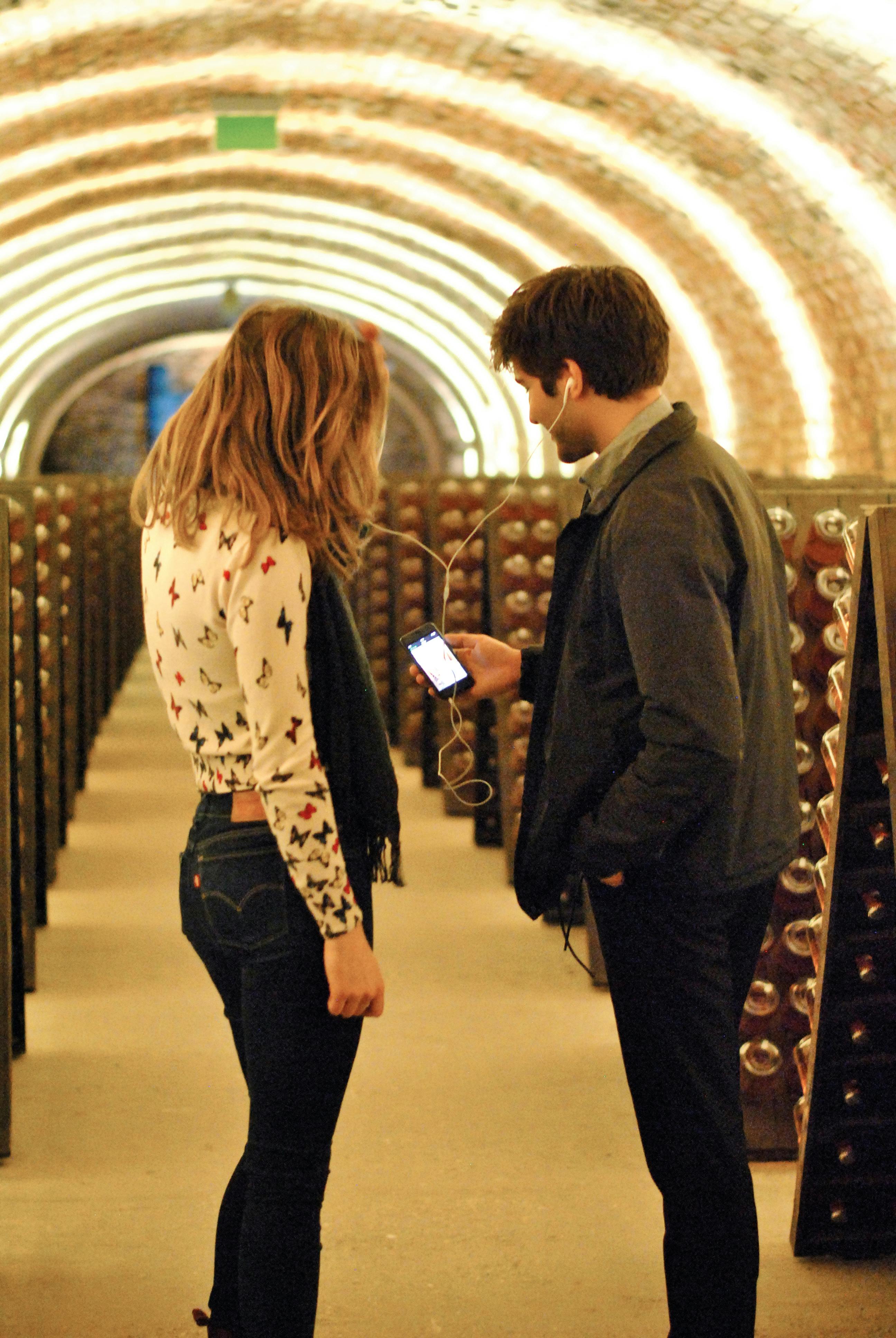 Two people, one wearing a white patterned shirt and the other in a dark jacket, share earphones while looking at a phone in a wine cellar.