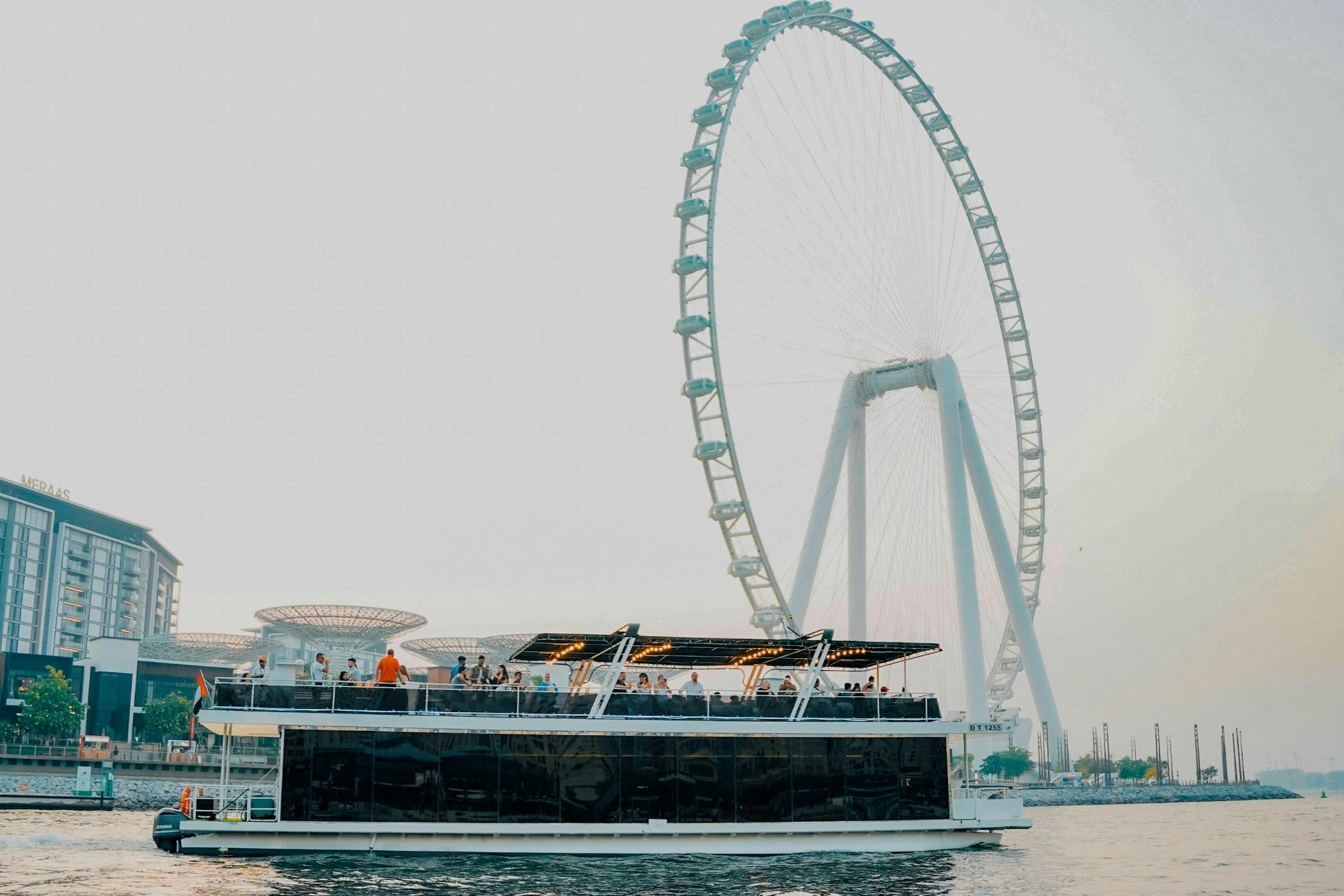 Лодка с большим количеством людей на верхней палубе плавает по воде рядом с большим колесом обозрения.