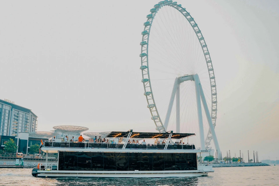 A boat with many people on the upper deck is floating on water near a large observation wheel structure.
