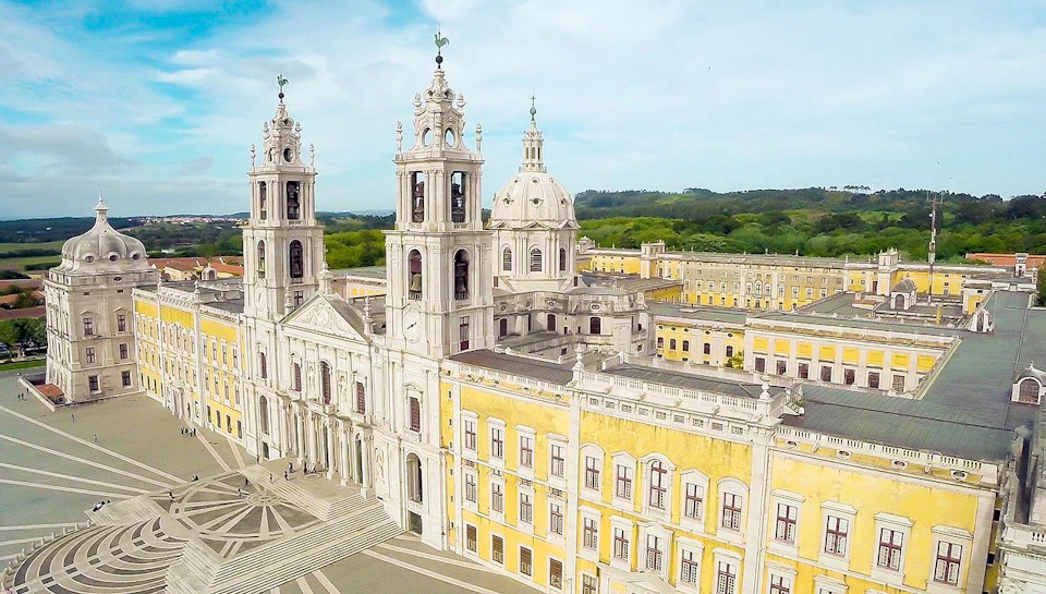 Aerial view of the Mafra National Palace, featuring twin towers, a dome, yellow walls, and a geometric stone courtyard.