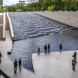 Besökare vid ett nivåindelat vattenfall med omgivande stentrappor och murar, beläget i ett urbant parkområde med träd och parkerade bilar.