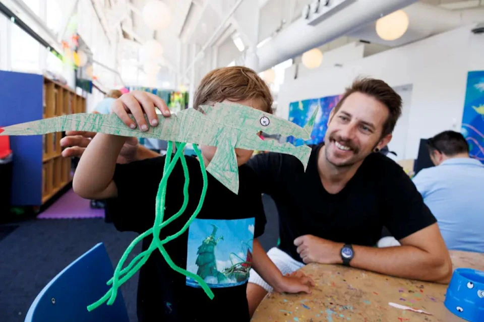 A child holds up a handmade paper craft resembling a crocodile while sitting next to a smiling adult in an art room.